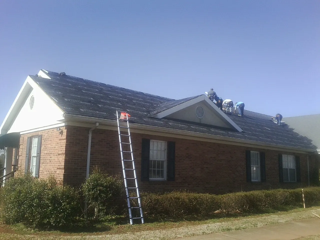 Crew tearing shingles off a brick ranch roof with a ladder set — Emergency Roof Repair work by our crew in Camp Dennison