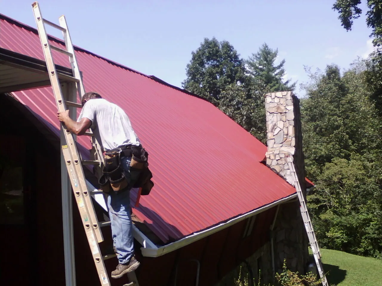 Roofer on a ladder beside a red metal roof and stone chimney — Soffit & Fascia Repair by Roofing Brothers in Camp Dennison